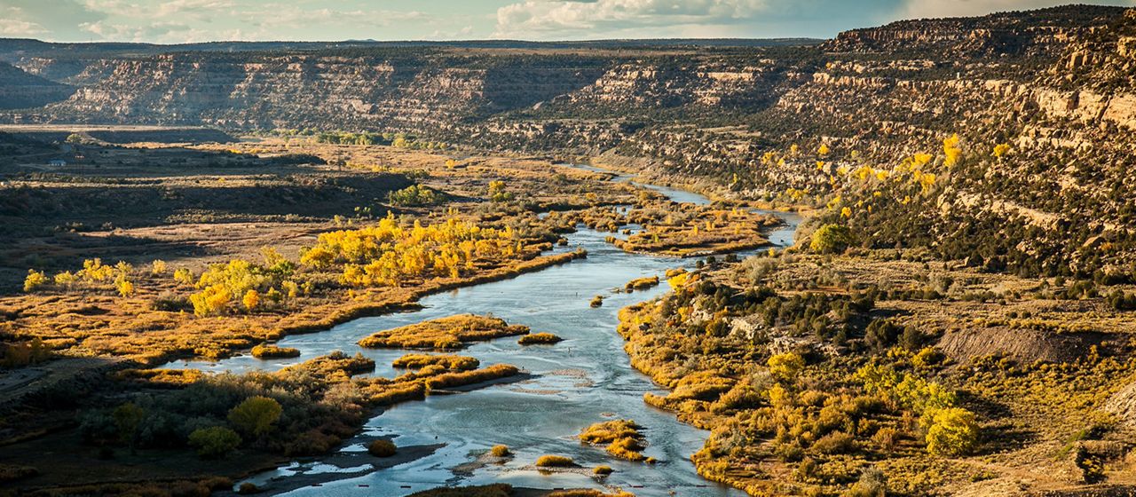 A wide river is running through a landscape with shrubs and mesas in the background.