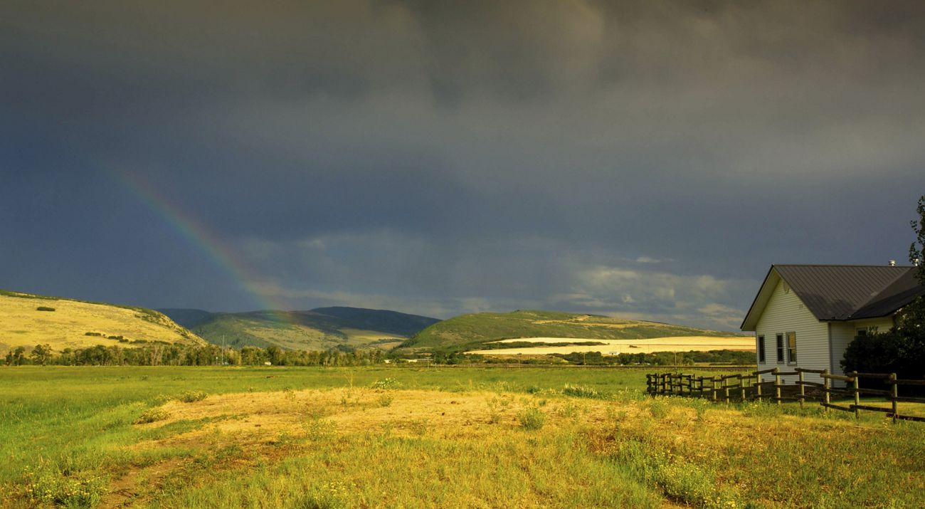 A house, wooden fence, fields and hills under a dark blue sky with a faint rainbow at Carpenter Ranch in Colorado.