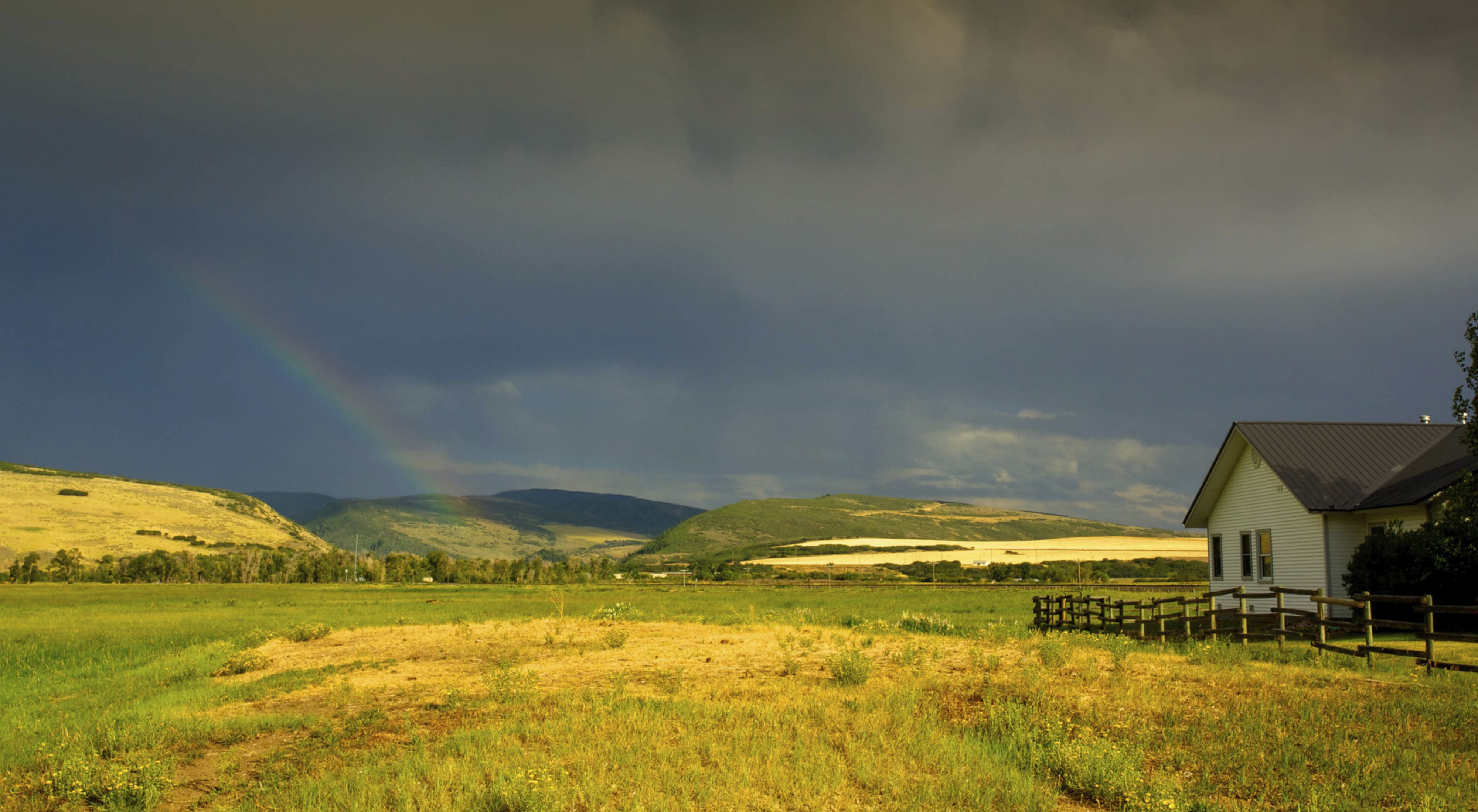 A house, wooden fence, fields and hills under a dark blue sky with a faint rainbow at Carpenter Ranch in Colorado.