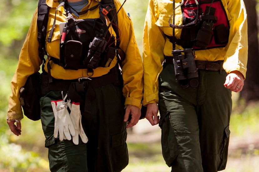 Two women wearing yellow fire retardant gear walk together down a wide dirt path in a TNC preserve during a controlled burn. They both wear red vests holding walkie-talkies.