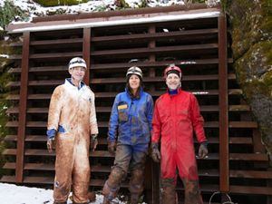 Three people stand together in front of a metal barrier at the opening to a cave. They wear coveralls covered in mud and ripped in places where they had to crawl through narrow cave openings.