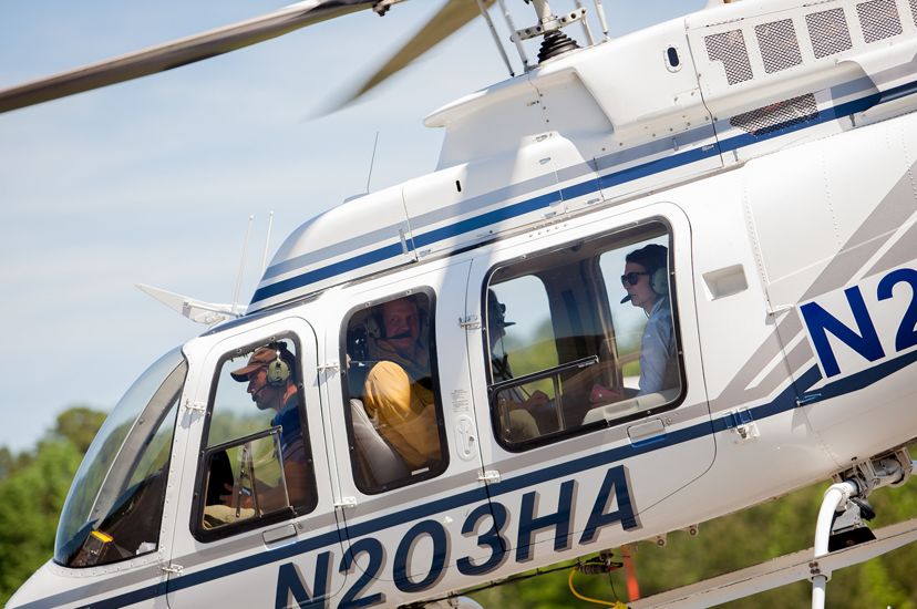 A white helicopter lifts off from a small airport. The pilot and three passengers are visible through the windows.