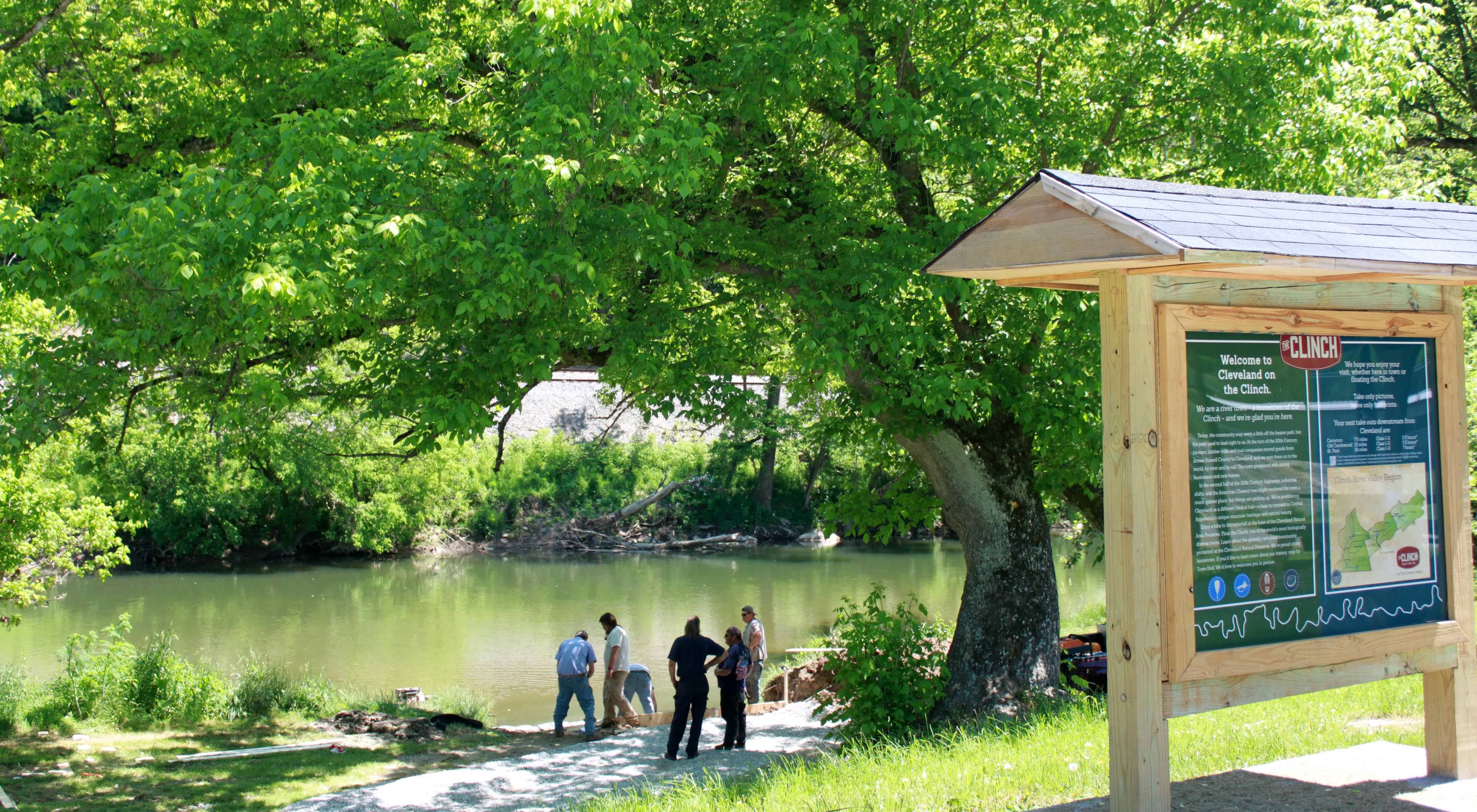 Six people stand at the edge of a tree lined river, inspecting an area where a canoe launch is being built. A large information kiosk in the foreground provides information about the river.