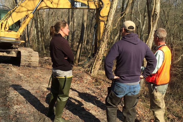 Three people stand near a yellow earth mover, a piece of heavy machinery used to cut breaches in the banks of the river behind them. The river is only barely visible through the trees.