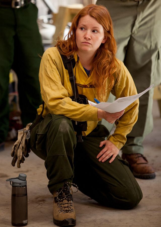 A woman wearing yellow fire retardant gear kneels on the floor during a briefing for a controlled burn.  