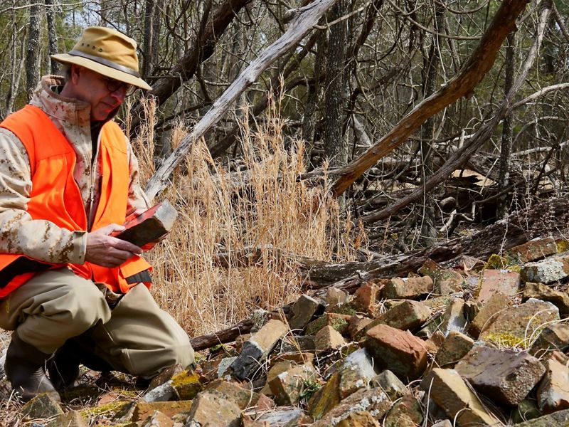 A man kneels next to a large pile of bricks that lay abandoned at the edge of a marsh. He holds one brick in his hands, examining it.
