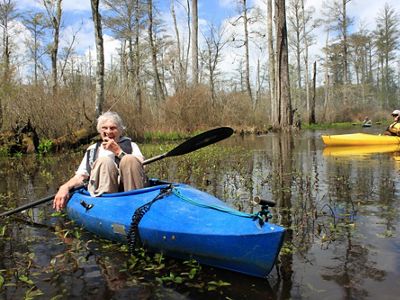 A smiling woman sits in a blue kayak floating in a cypress swamp. A man in a yellow kayak paddles behind her.