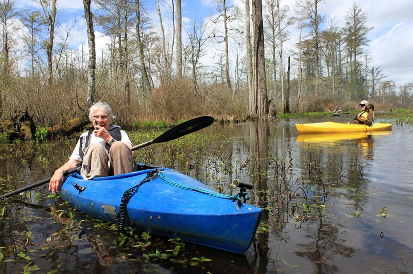 A woman sits in a blue kayak holding a small microphone to her mouth as she guides a tour along the wide, flat water of Dragon Run. A man in a yellow kayak floats behind her.