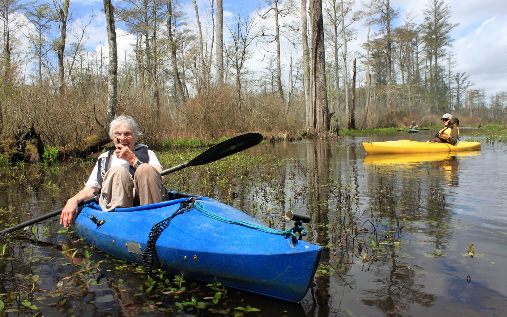 A smiling woman sits with her knees drawn up in front of her in a blue kayak. Green submerged vegetation floats beneath her. A man in a yellow kayak is behind her.