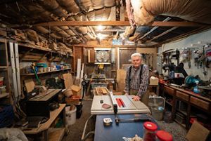 A man standing in a wood shop with wood and tools surrounding him. 