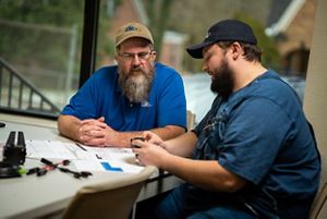 Two men sit at a table looking over sheets of papers and blueprints. 