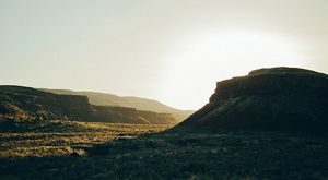 The face of a mesa sits in darkness as the sun glows behind it, filling the desert valley with light.