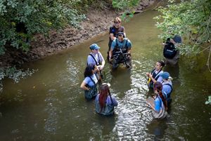A group of people wading in a river while a cameraman films them. 