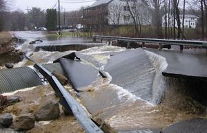 A washed out culvert overtopped by floodwaters and a broken up road.