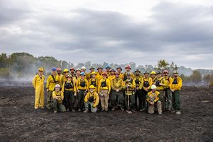 A group of about 20 people wearing bright yellow protective gear is gathered at a smoky barrens under a cloudy, gray sky.