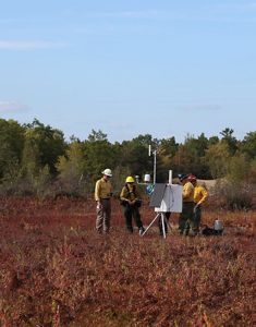 A small group of people in yellow and white protective gear are huddled around a pop-up whiteboard in the middle of a barrens; below them are red grasses, and above them is a bright blue sky.
