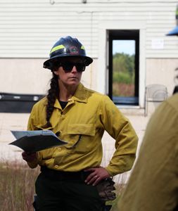 A woman in yellow protective gear and sunglasses is addressing a mentee in front of a plain concrete building.
