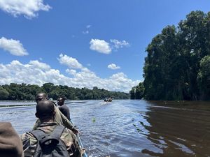 Men in a boat head down a river lined with trees on a sunny day.