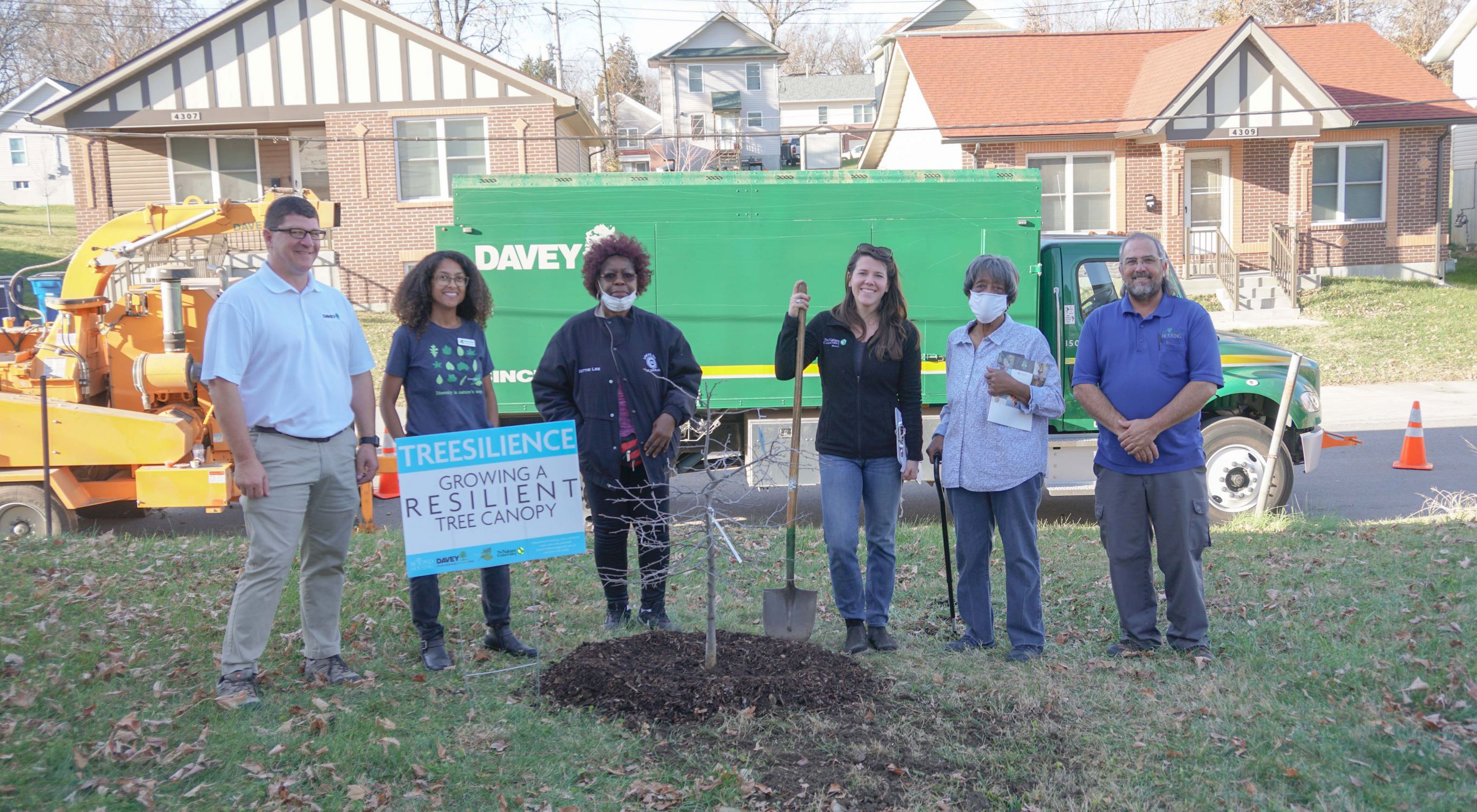 A small group of adults stand by a newly planted tree in an urban back yard.