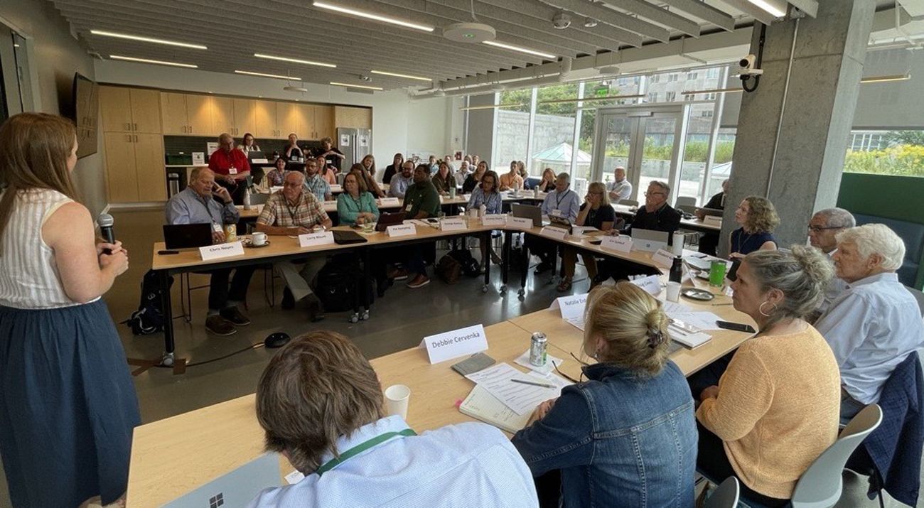 A group of people sitting around long tables arranged in U shape listening to a person talking at the left of the image.