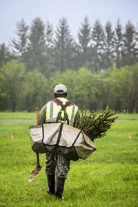 The back view of a man walking with a pickaxe and a bag full of tree saplings.