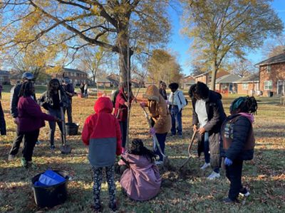 A group of girls gather together to plant a tree sapling. Two girls stabilize the tree while other girls fill in the hole with dirt.