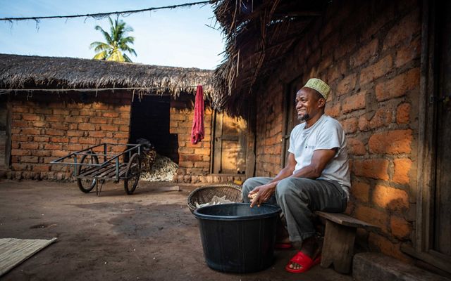 man sitting with bucket of maize in front of small brick home