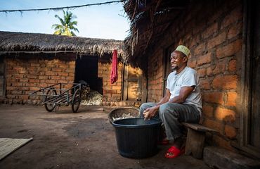 man sitting with bucket of maize in front of small brick home
