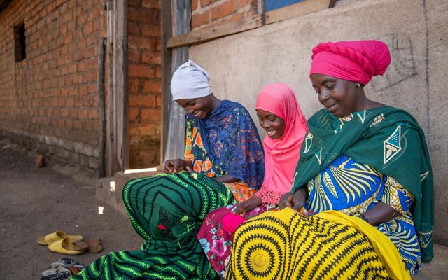 A woman and two daughters embroider blankets as they sit beside their house.