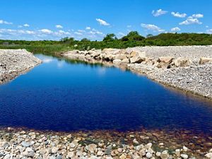 A narrow channel of open water reflects the clear blue sky, with heaping piles of small stones forming both banks and green dune vegetation in the background