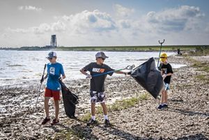 Three young boys in baseball caps use grabbers to pick up trash along the water's edge at Texas City Prairie Preserve.