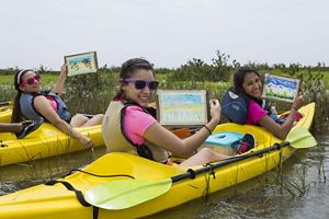 Three young girls sit in bright yellow kayaks, showing off their paintings of nature.