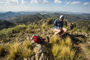 A woman sits at the top of a rocky mountain covered in grass as numerous mountains rise and fall in the background.