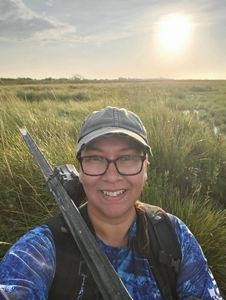 A person wearing a hat and glasses poses in front of a grassy field.