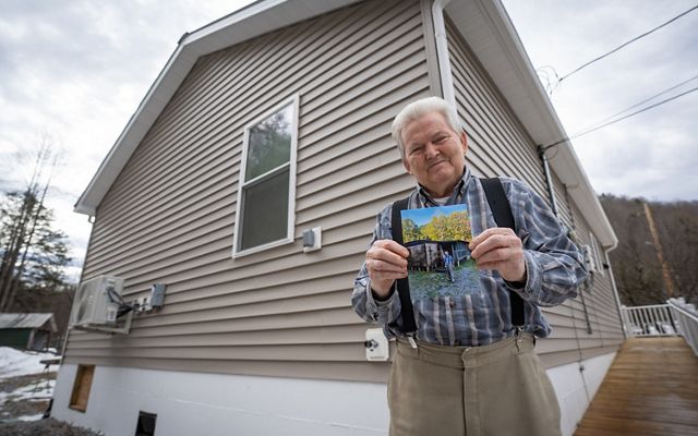 A man smiling and holding a photo of his old home in front of his new, energy efficient house. 