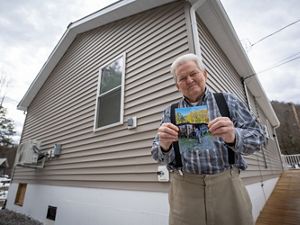 Image of an older man smiling and holding a photo of his old house which was lost in a flood while posing in front of his new raised home. 