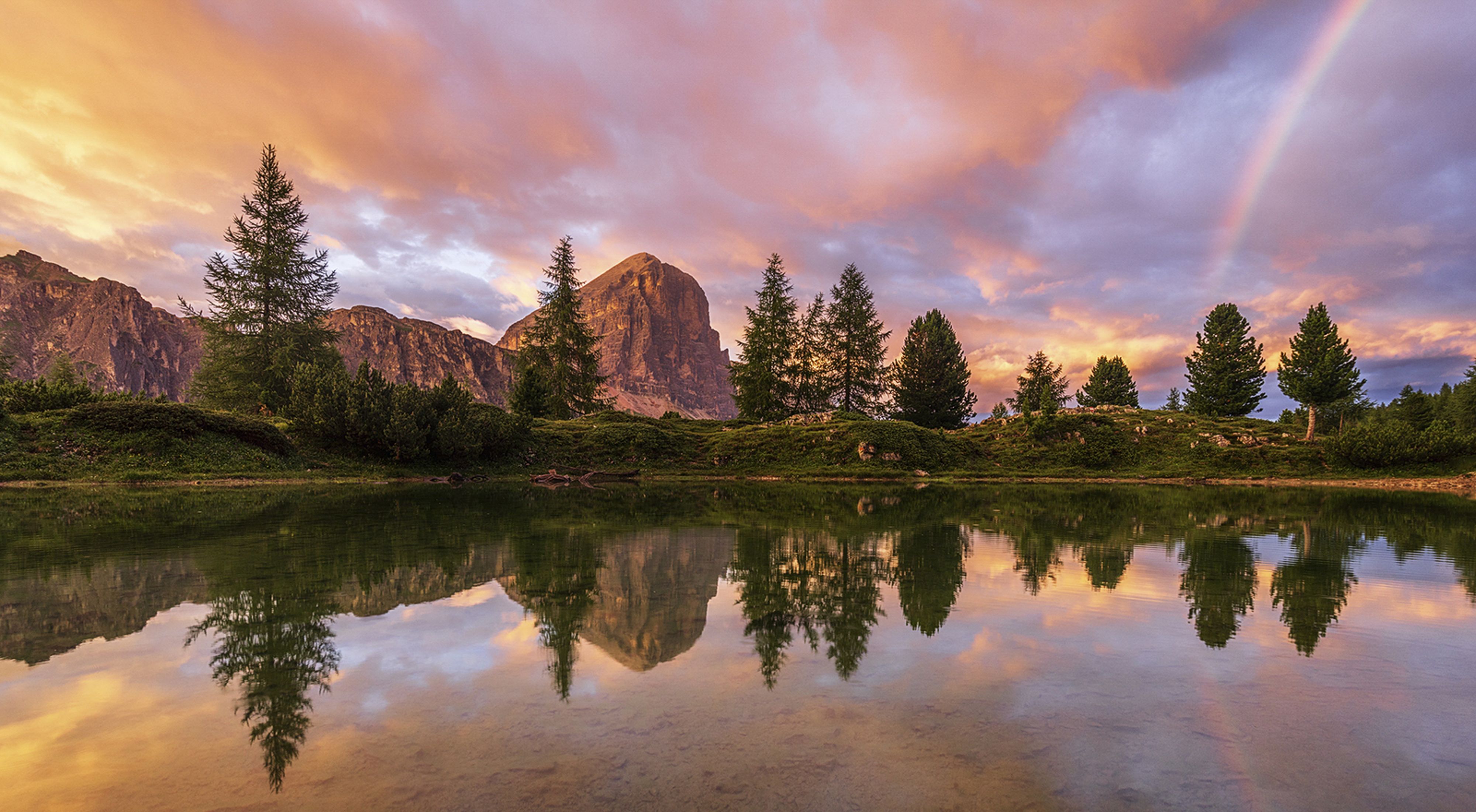 sparse evergreen trees and rocky outcroppings and a rainbow reflected in a still lake at sunset
