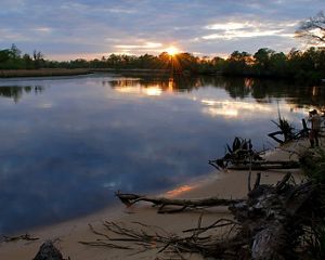 The sun sets over a placid body of water as a person stands on the water's banks and takes a photo.