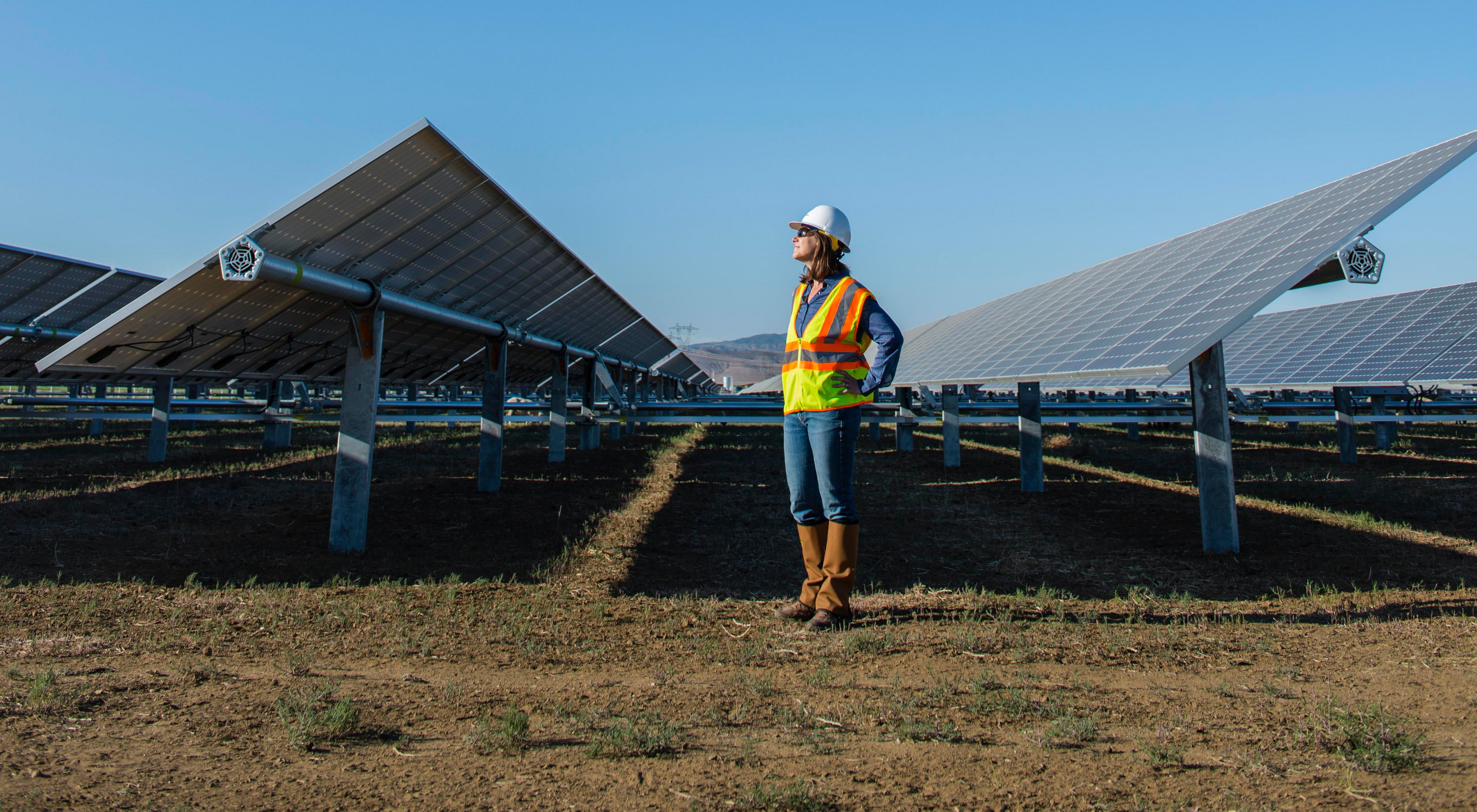 A woman stands on the ground in between two solar arrays wearing a reflective orange vest and a construction hard hat, looking to the upper lefthand side of the screen.
