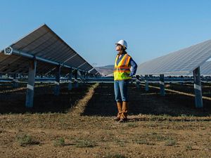 A woman stands on the ground in between two solar arrays wearing a reflective orange vest and a construction hard hat, looking to the upper lefthand side of the screen.