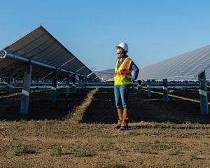 A woman stands on the ground in between two solar arrays wearing a reflective orange vest and a construction hard hat, looking to the upper lefthand side of the screen.