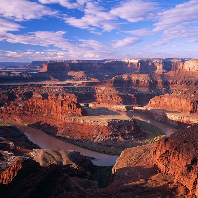 A wide river curves through a landscape of towering mesas that glow red and golden in the sun.