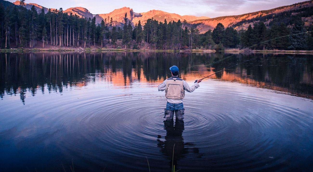 A person wearing a vest and a blue hat stands in the middle of a lake and casts a fishing line while the calm water ripples around him.