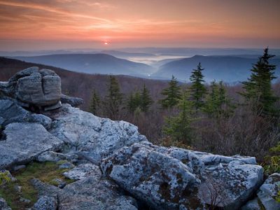 Sunrise over a vast expanse of forested mountains with large boulders in the foreground.