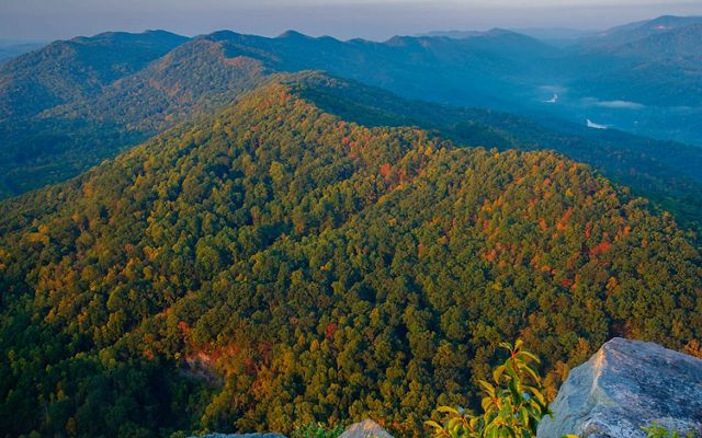 Aerial view looking out over a mountain ridge that extends into the distance where it meets another ridge at the horizon. The trees on its steep forested flanks are just beginning to show fall colors.