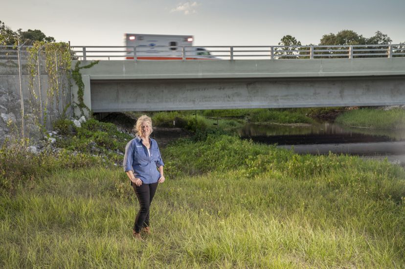 woman stands on grassy riverbank under a highway as an ambulance drives by