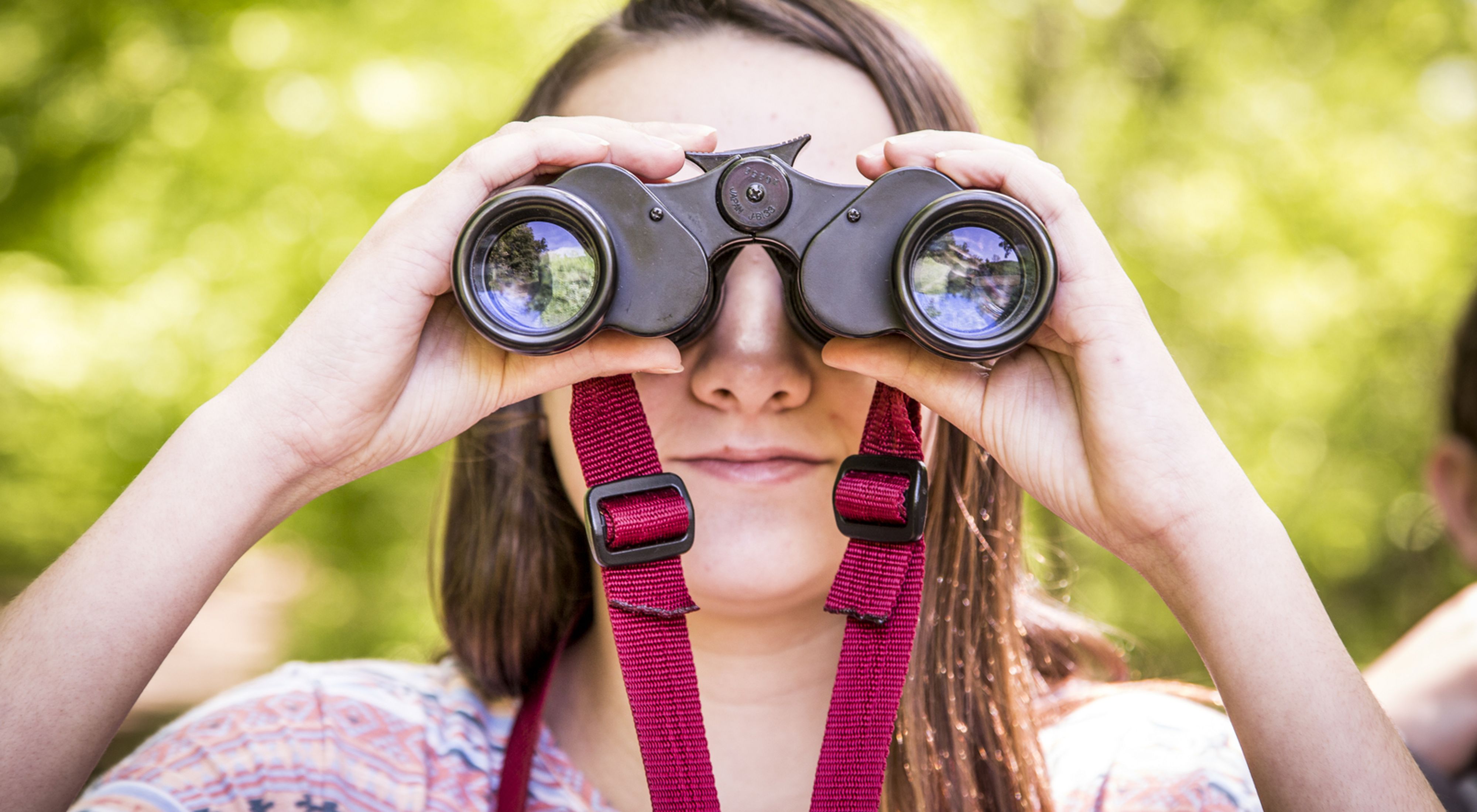 A close-up photo of a person looking at the camera through binoculars.