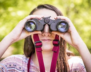 A close-up photo of a person looking at the camera through binoculars.