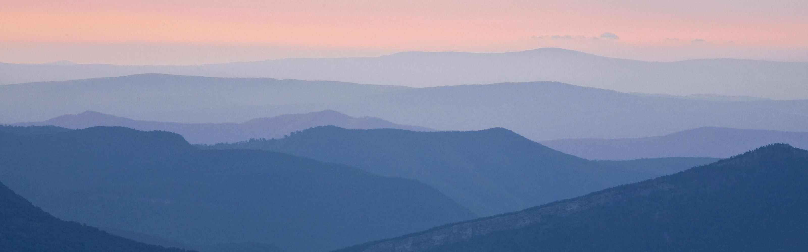 View looking across a vast series of mountains, each more pale in color as they reach toward the horizon, under a dusky orange and purple sunrise sky.