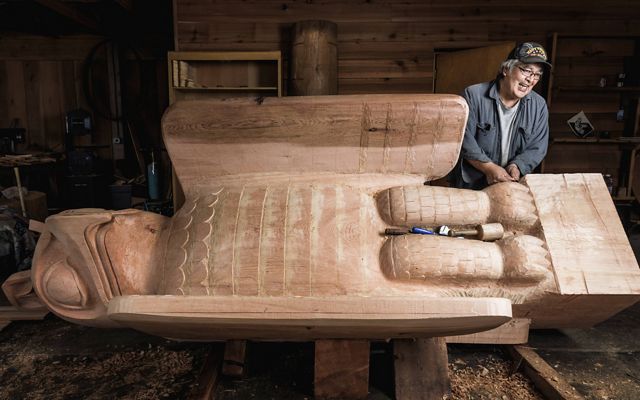 Within a carving shed, a man leans on a freshly carved totem pole of a sea wolf, made out of cedar wood